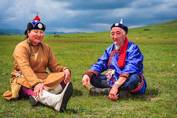 Two people sit on a wide green grassland beneath a dramatic, cloudy sky. The man wears a bright blue robe with a red scarf, a traditional hat topped with a small ornament, prayer beads and worn boots; the woman wears a tan robe, white embroidered boots and a decorated hat, smiling gently. Rolling hills stretch into the background, creating a peaceful, open-steppe scene that highlights traditional clothing and relaxed conversation outdoors.