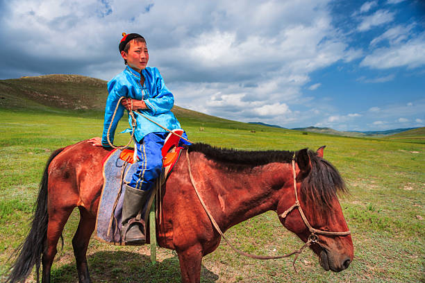 A young rider wearing a bright blue traditional coat and hat sits astride a chestnut horse in a wide, grassy valley. He holds the reins and rests one hand on the horse’s back while the animal grazes with its head lowered. Rolling green hills stretch into the distance under a dramatic partly cloudy sky, creating a vivid, pastoral scene.