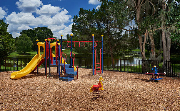 A bright and colorful playground with slides, climbing bars, and spring toys sits on a bed of wood chips near a pond and trees under a sunny blue sky with scattered clouds.