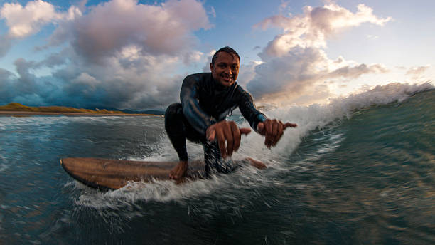 Close-up view of young man surfing wave at sunrise, lofty, pink clouds distant, Kuril Islands