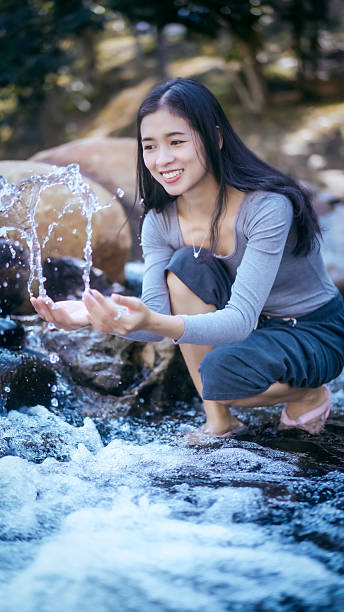 Young woman crouching by a rocky stream outdoors, smiling and catching splashing water with hands, natural forest background, soft light, lifestyle nature concept