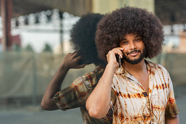 Smiling man with afro hair talking on smartphone next to glass wall with reflection
