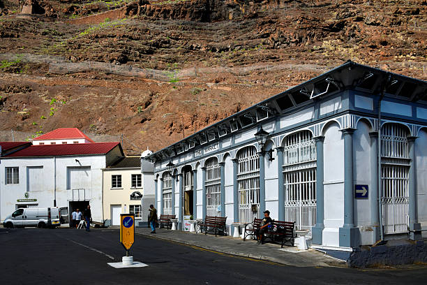 Jamestown, Saint Helena: the historic Market building located on Market Street - constructed in 1865 using prefabricated cast iron to resist termite damage, the structure features a central clock, arched entryways, and repetitive architectural detailing characteristic of Victorian-era civic engineering. The building serves as a retail hub for the isolated British Overseas Territory, accommodating local agricultural vendors and small businesses. The surrounding urban environment includes adjacent commercial structures and the clock tower. Rising steeply behind the settlement are the barren volcanic cliffs that define the deep, narrow ravine of Jamestown.