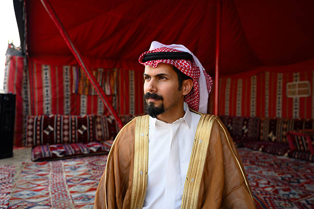 Middle Eastern man looking away, dressed in a traditional ghutra, thobe, and bisht, sitting thoughtfully inside a vibrant desert tent, representing Arabian culture and heritage
