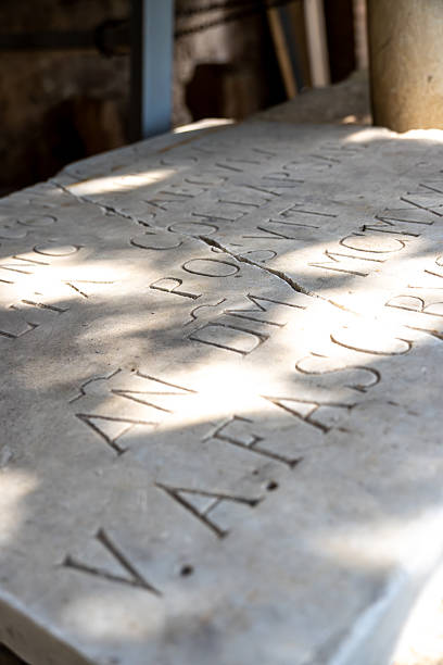 Vieste - Apulia - Italy - 08-29-2025: Low angle close-up shows carved Latin letters on a cracked stone slab, with sunlight, leaf shadows, and blurred columns behind it.
