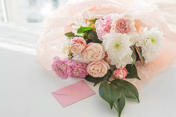 Delicate bouquet of blush roses pink carnations and white chrysanthemums wrapped in soft paper with pastel envelope and heart symbol, airy daylight background, romantic greeting concept for holiday branding and celebration design