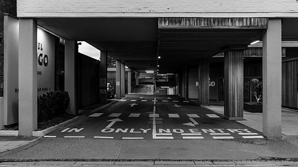 Black and white photo of a covered driveway with directional arrows and 'in only' marking, flanked by columns and storefronts, leading to a parking garage entrance