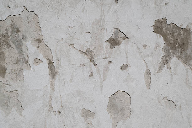 Close-up of a damaged white plaster wall with peeling paint, cracks and exposed concrete underneath. The worn surface and irregular patches create a raw, textured background suitable for themes of decay, aging and renovation.
