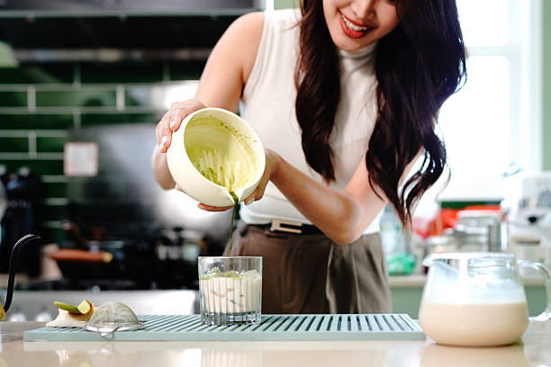 Smiling asian woman preparing iced matcha latte by pouring whisked green tea over milk at modern kitchen counter, preparing traditional Japanese green tea