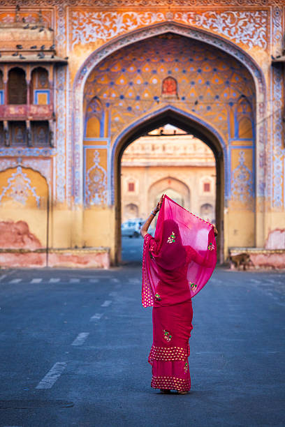 A person seen from behind lifts a sheer magenta veil while standing in the middle of an empty street, framed by a large, ornately painted arched gateway. The façade displays intricate floral and geometric motifs in warm ochres, blues and pastels, creating a vivid backdrop that contrasts with the deep pink of the traditional dress. The composition emphasizes the arch framing and leading lines of the road, with small details—pigeons on the structure and weathered plaster—adding to the scene's lived, historic character.