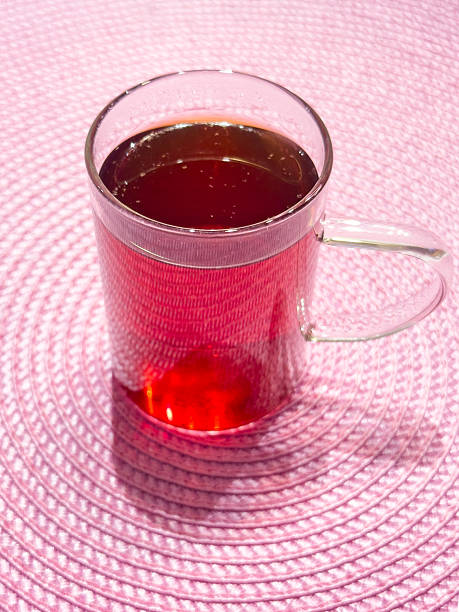 A clear glass teacup filled with steaming hot reddish tea stands elegantly on a textured pink woven placemat, casting soft shadows that enhance the warm inviting glow. This simple yet sophisticated beverage presentation conveys comfort and tradition, making it suitable for tea shop displays, relaxation campaigns or social media tea appreciation posts. Horizontal composition, high resolution, taken on mobile device, suitable for commercial use.