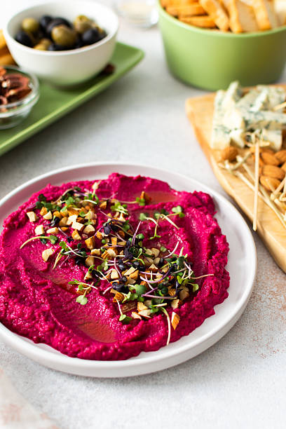 Beetroot hummus is placed in the center of a charcuterie board, surrounded by snacks like olives, nuts, cheese, and crispy breadsticks. A light, inviting setup showcases vibrant colors