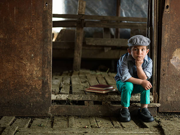 Full length photo of well dressed 6 years old boy posing in barn. He is wearing a newsboy cap, gray shirt and waist along with green pants. Shot with a medium format camera under natural light.