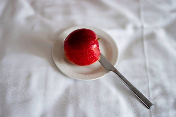 Red apple on a small white plate with a knife resting on soft white bedding, simple and fresh.