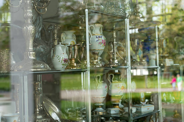 Antique porcelain vases and silver candlesticks displayed in vintage shop window with soft reflections of green park outside.