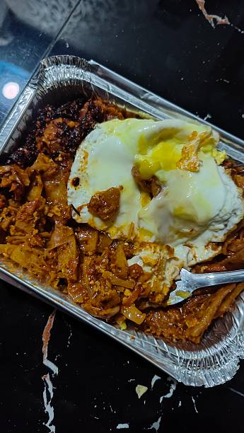 A savory and spicy take-out meal featuring seasoned noodles topped with a sunny-side-up fried egg in an aluminum foil container.