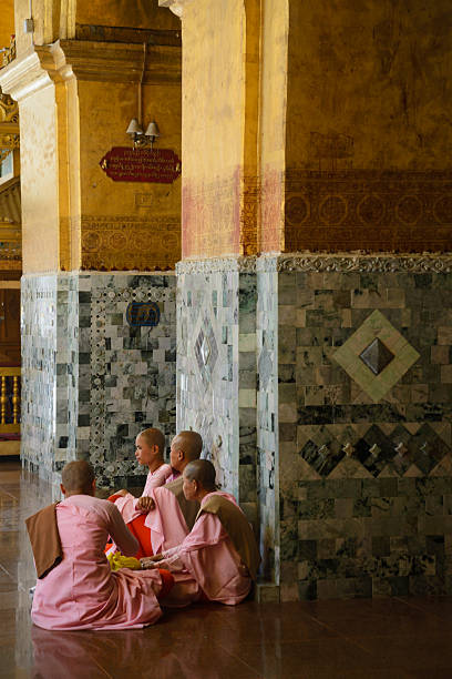 A group of 4 nuns (thilashin) with the traditional pink and orange uniform and shaved head, sitting on the floor of the temple coumpound