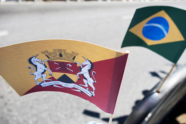 A vibrant Sorocaba flag, with its unique coat of arms, is prominent in close-up. The Brazilian flag is blurred behind. Bright sunlight creates sharp shadows on the pavement, showcasing civic pride.