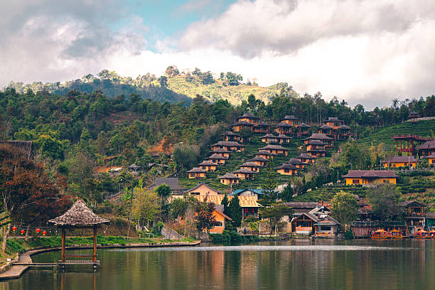 A scenic view of a Chinese-style accommodation on a hillside amidst tea plantations in Ban Rak Thai, Mae Hong Son, Thailand. A large reservoir stands in the foreground, its reflection visible in the water. There is a wooden pavilion in the middle of the water and a walkway along the reservoir.