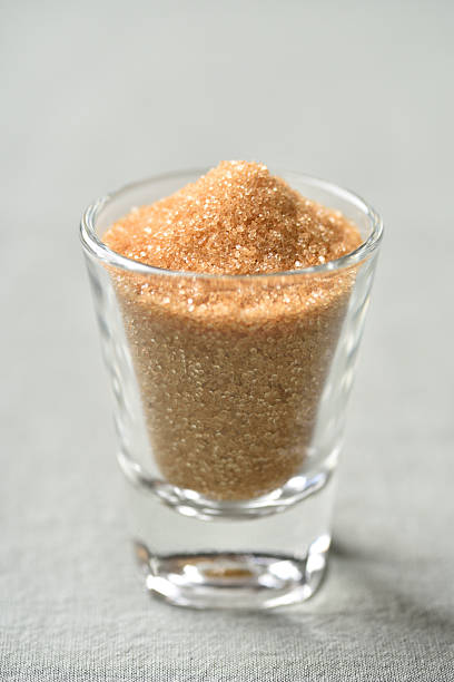 Organic brown sugar in a small clear glass bowl placed on a green fabric background. Studio shot of natural unrefined sugar for cooking and baking
