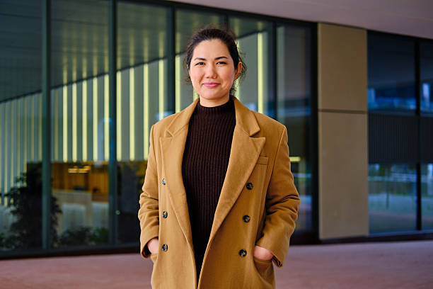 Asian woman posing confidently, hands in pockets, wearing an elegant coat in a city setting