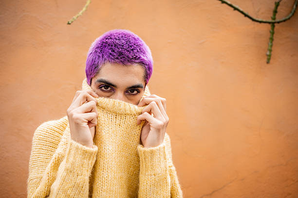Young adult with purple buzz cut hair hiding lower face with a yellow knitted sweater, looking at camera