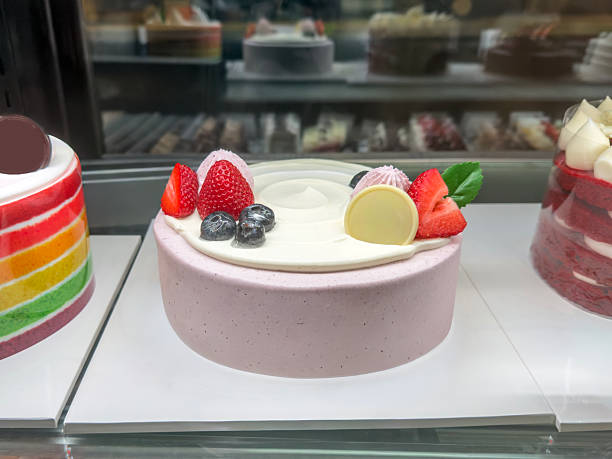 Full frame view of elegant white cream Strawberry Soft cake topped with fresh strawberries white chocolate disc and blueberries on blue rimmed base labeled in glass fronted refrigerated pastry display case alongside pink berry cream cakes chocolate dome cake and cream tarts under store lighting with copy space. Luxurious bakery assortment showcases refined Japanese style confections ideal for dessert menu cafe promotion or gourmet patisserie concepts. Horizontal composition highlights variety and freshness for commercial food stock.​