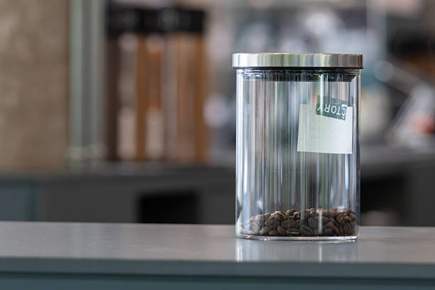 Glass jar holds dark coffee beans. Background shows blurred kitchen shelves