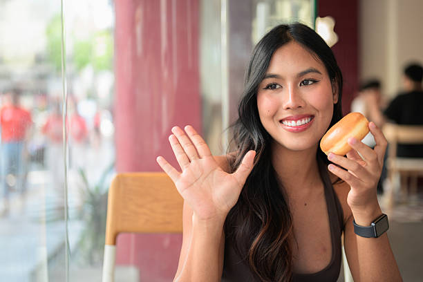 Young woman sitting in a cafe holding a glazed donut while waving her hand in greeting