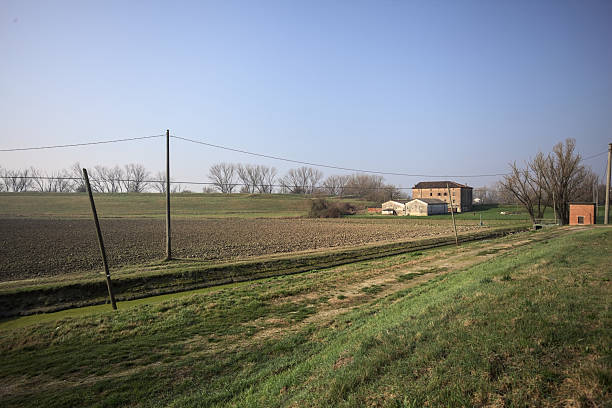 Country house with barns, on a plowed field, with an embankment at the edge of the field, and a trench with water, with a small dam on it, on a sunny day in the italian countryside