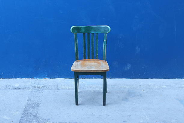 A vintage, weathered wooden chair stands as a simple focal point against a vibrant, solid blue wall. The chair features a dark, teal-painted frame with visible signs of age and wear, contrasting with its light, natural-finished wooden seat. Positioned centrally on a textured concrete floor, the scene emphasizes a minimalist, rustic aesthetic through its stark composition and bold use of color.