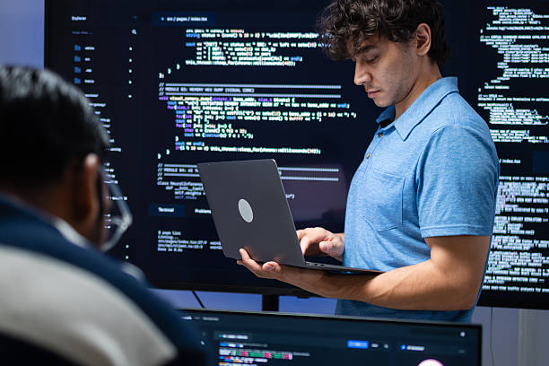 Programmer Working with Laptop and Code Display, Reviewing Software Tasks While Standing in Front of a Large Programming Screen in Modern Tech Office.