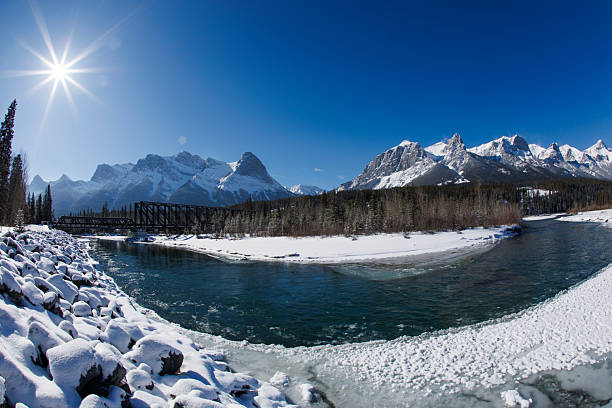 A view of the Bow River and the old Canmore Engine Bridge in Alberta, Canada, in the wintertime. The Canmore Engine Bridge is an iconic landmark in the Canadian Rockies and now serves as a pedestrian bridge that crosses the Bow River. From the left, the Three Sisters mountains, Ha Ling Peak, and Mt. Rundle are visible.
