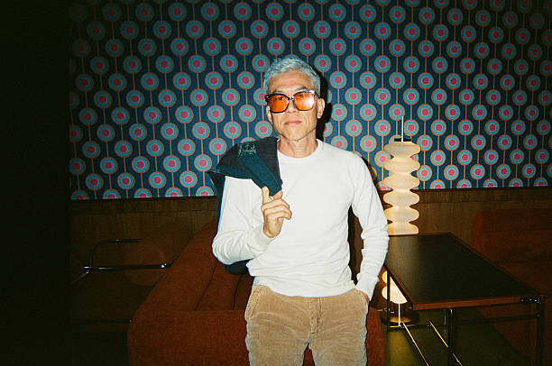 Stylish mature Asian man wearing tinted glasses and holding a jacket while posing casually in a retro lounge setting with instant film aesthetic, Kuala Lumpur