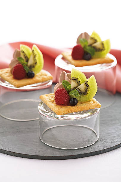 Crispy crackers topped with whipped cream, fresh kiwi slices, raspberries, blueberries, and mint leaves, served on overturned glass cups.