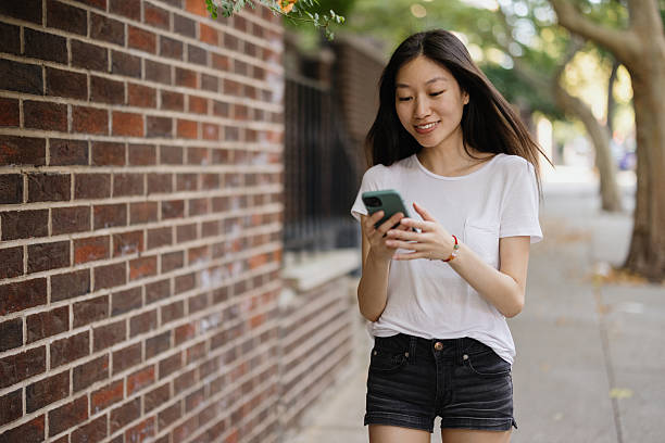 Young adult Asian woman walking on city street, smiling while looking at smartphone. Brick wall and trees in background