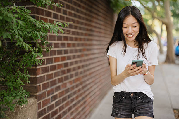 Smiling young adult asian woman walking confidently on a sidewalk, checking messages on her mobile phone