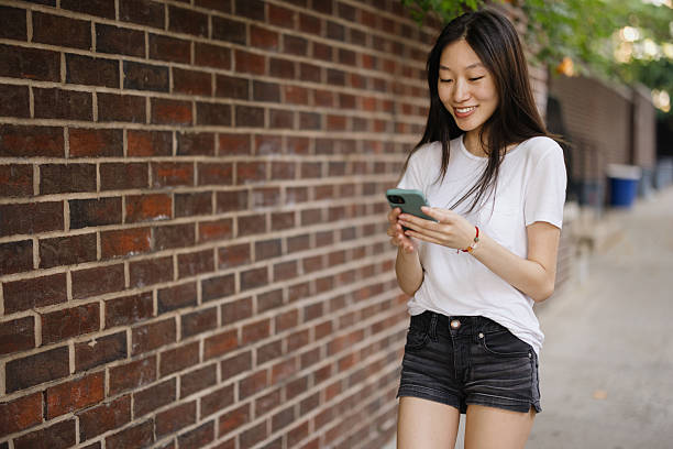 Young adult Asian woman smiling while messaging on her phone, standing casually outside a building with a brick wall
