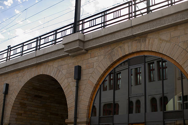 Arched viaduct with rail lines and window reflection, iron railing above stone arches, overhead wires crossing pale sky, rhythmic geometry suggesting