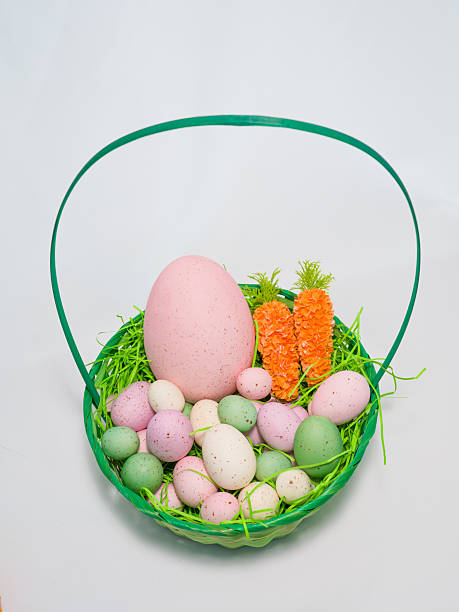 Green wicker basket filled with colorful speckled Easter eggs and decorative carrots on a white background.