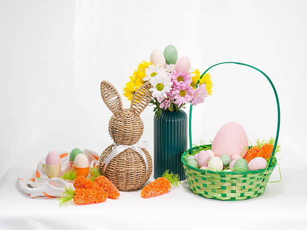 Festive Easter composition featuring a woven bunny, a basket filled with colorful eggs and carrots, and a floral bouquet on a white table.