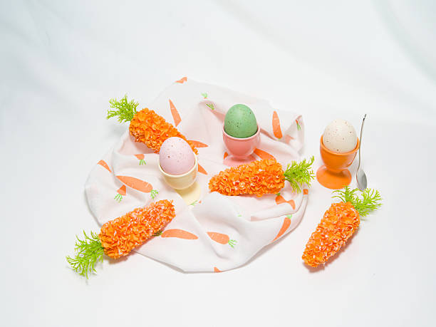 Three colorful speckled Easter eggs in ceramic cups with decorative carrots arranged on a patterned napkin.