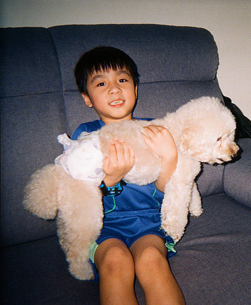 Young Asian boy sitting on a sofa while holding a fluffy pet dog, representing childhood companionship, family life, and the bond between children and pets, Kuala Lumpur