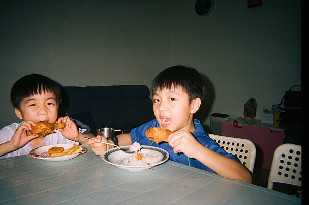 Two Asian boys eating fried chicken and rice at a dining table in a home setting, showing casual family mealtime, childhood appetite, and everyday domestic life, Kuala Lumpur