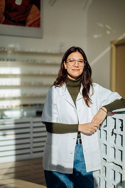 Optometrist wearing glasses and lab coat smiles in a bright optical shop with shelves of eyewear