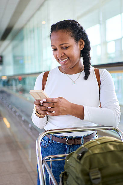 Vertical. African millennial woman checking her smartphone while pushing a luggage trolley in a bright airport terminal. Glass walkway and signage suggest boarding time, travel mood and anticipation.
