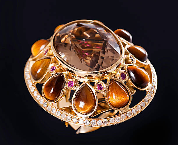 Details: Large ring with a faceted smoky topaz as the center stone, surrounded by the following stones: round pink sapphires, arranged between teardrop-shaped tiger's eye stones in shades of brown and brownish-orange; stones on the edges; a row of small diamonds along the lower edge, black background.