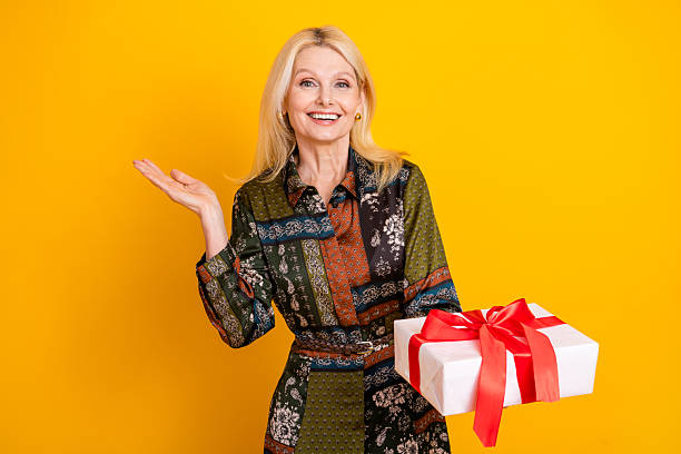 A stylish mature woman with blonde hair stands against a vibrant yellow backdrop, smiling as she offers a wrapped gift with red ribbon for a celebratory promotional feel.
