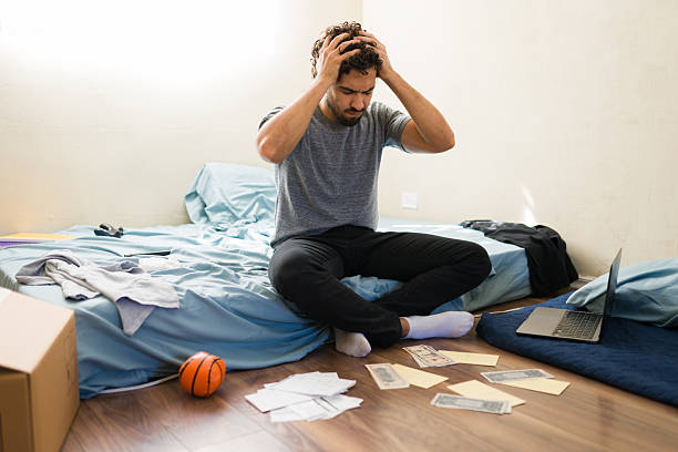 Young man holding head in despair, sitting on a bed in a messy room, counting bills and struggling with financial stress