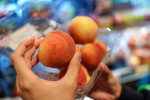 A close-up view of a person's hands holding an open transparent plastic container filled with fresh, ripe peaches in a supermarket retail setting.